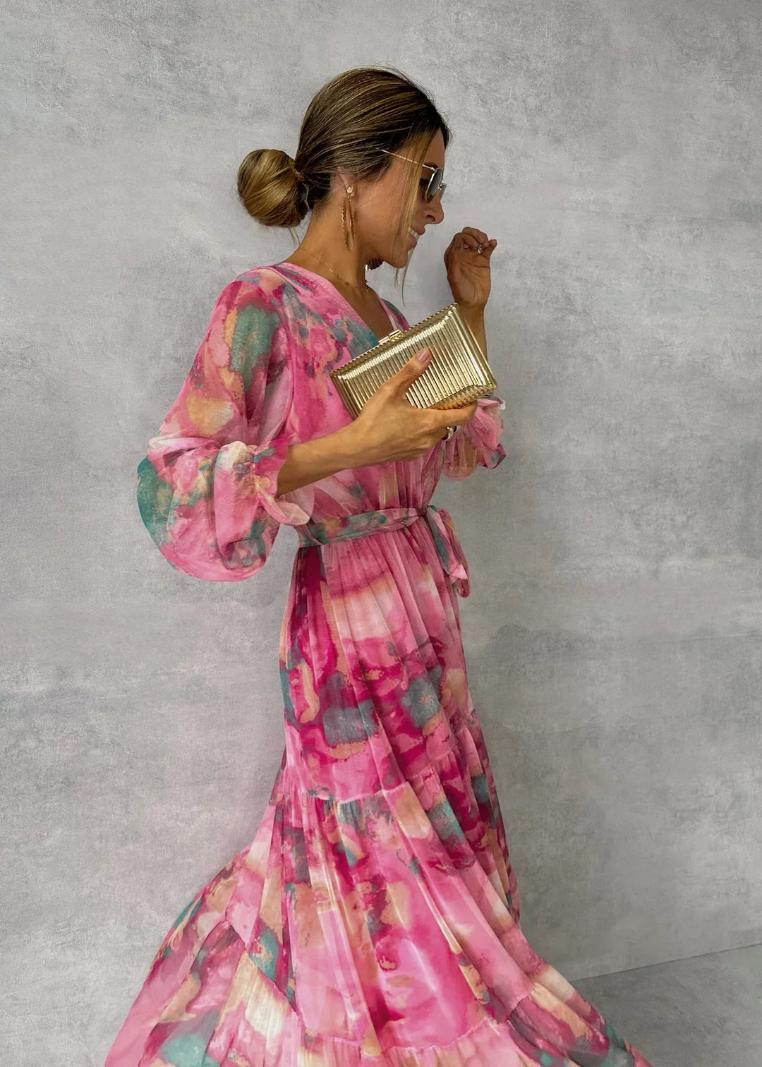 Frau in einem eleganten, rosa-blumigen Maxikleid mit goldener Clutch und Sonnenbrille vor grauem Hintergrund. Fashion, Damenmode, Abendkleid.