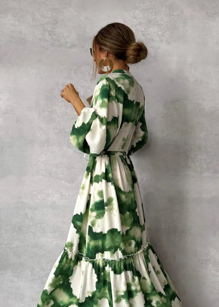 Frau in grün-weißem Batik-Maxikleid mit langen Ärmeln und Taillengürtel, seitlich stehend vor grauer Wand, eleganter Look, Mode 2023.