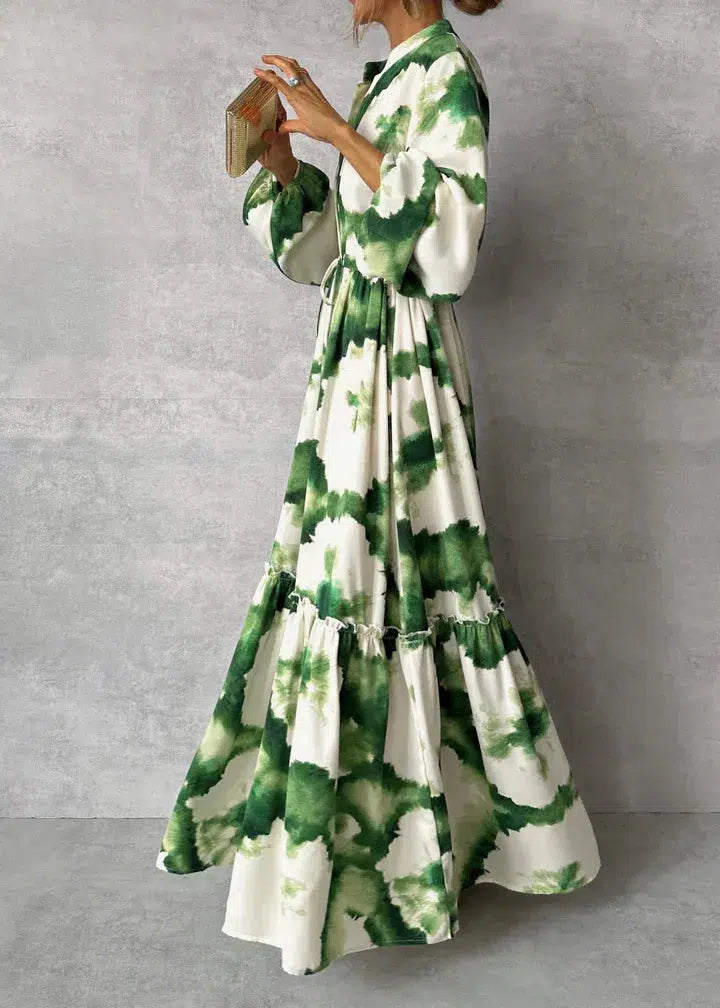 Frau in langem, grün-weißem Batik-Kleid mit Puffärmeln und Gürtel, hält eine kleine Tasche. Elegantes, modernes Mode-Outfit für besondere Anlässe.