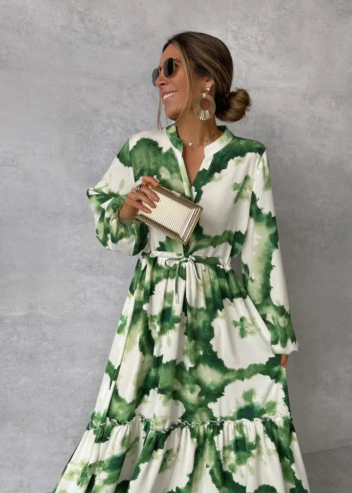 Frau in grün-weißem Blumenkleid mit Sonnenbrille und goldener Clutch vor grauem Hintergrund. Elegante Mode, Sommerkleid, Accessoires.