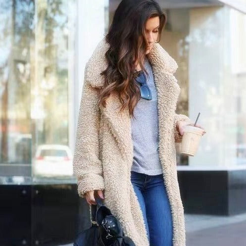 Frau in beigem Teddy-Mantel, grauem Shirt und blauer Jeans, hält Eiskaffee und schwarze Tasche, spaziert in städtischer Umgebung.