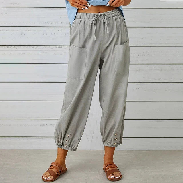 Graue Damenhose mit Taschen und Knöpfen am Saum, kombiniert mit braunen Sandalen. Bequeme Sommermode vor weißer Holzwand.