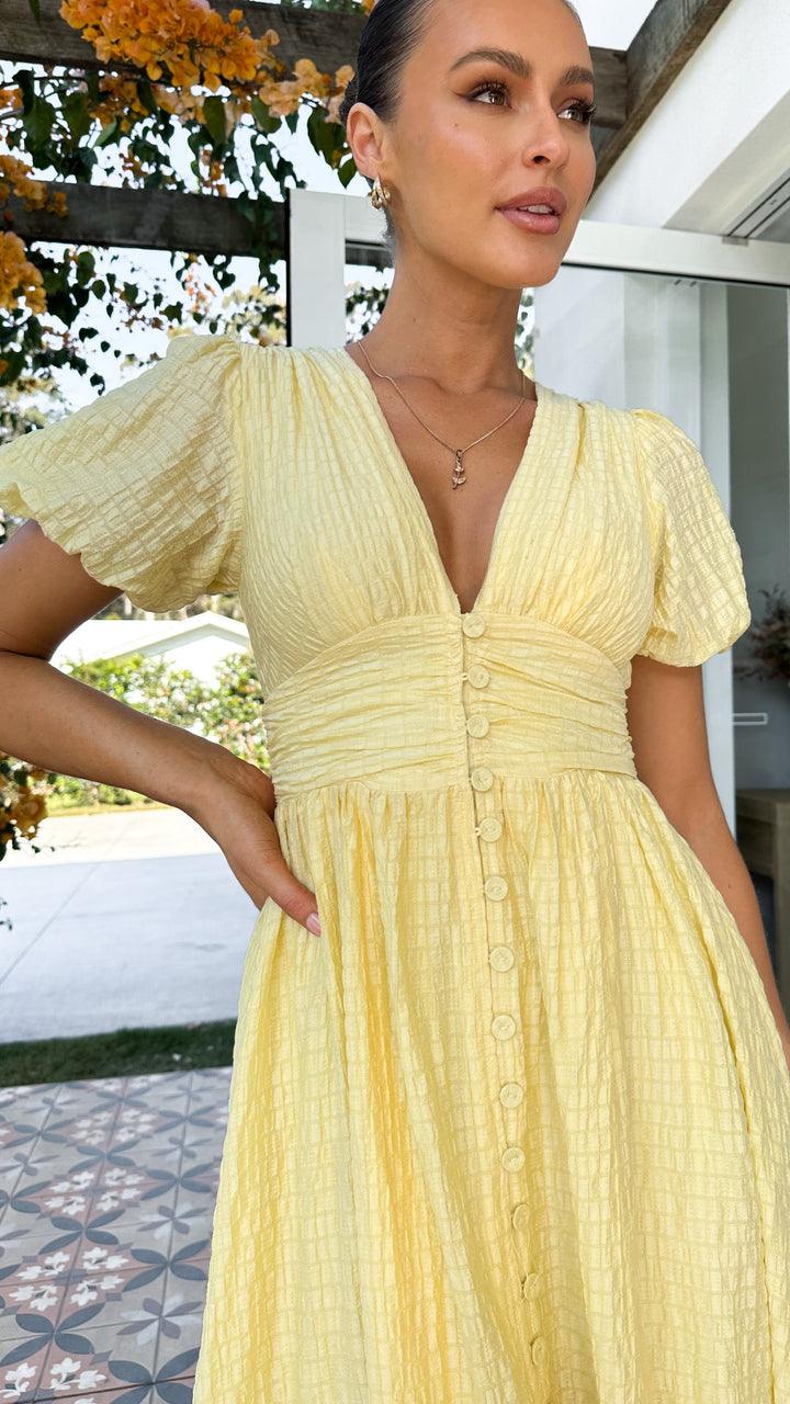 Frau in gelbem Sommerkleid mit Knopfleiste, steht draußen vor modernem Gebäude, umgeben von blühenden Pflanzen. Elegantes, luftiges Design.