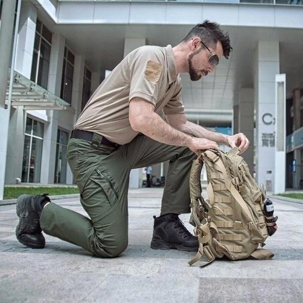Mann in taktischer Kleidung kniet und überprüft einen beigen Rucksack vor einem modernen Gebäude. Outdoor-Ausrüstung, taktische Mode, urbane Umgebung.