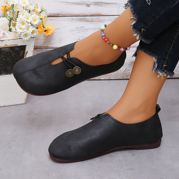 Schwarze Damen-Lederslipper mit dekorativen Knöpfen, getragen mit Jeans und buntem Fußkettchen, neben einem Blumenkasten auf grauem Boden.