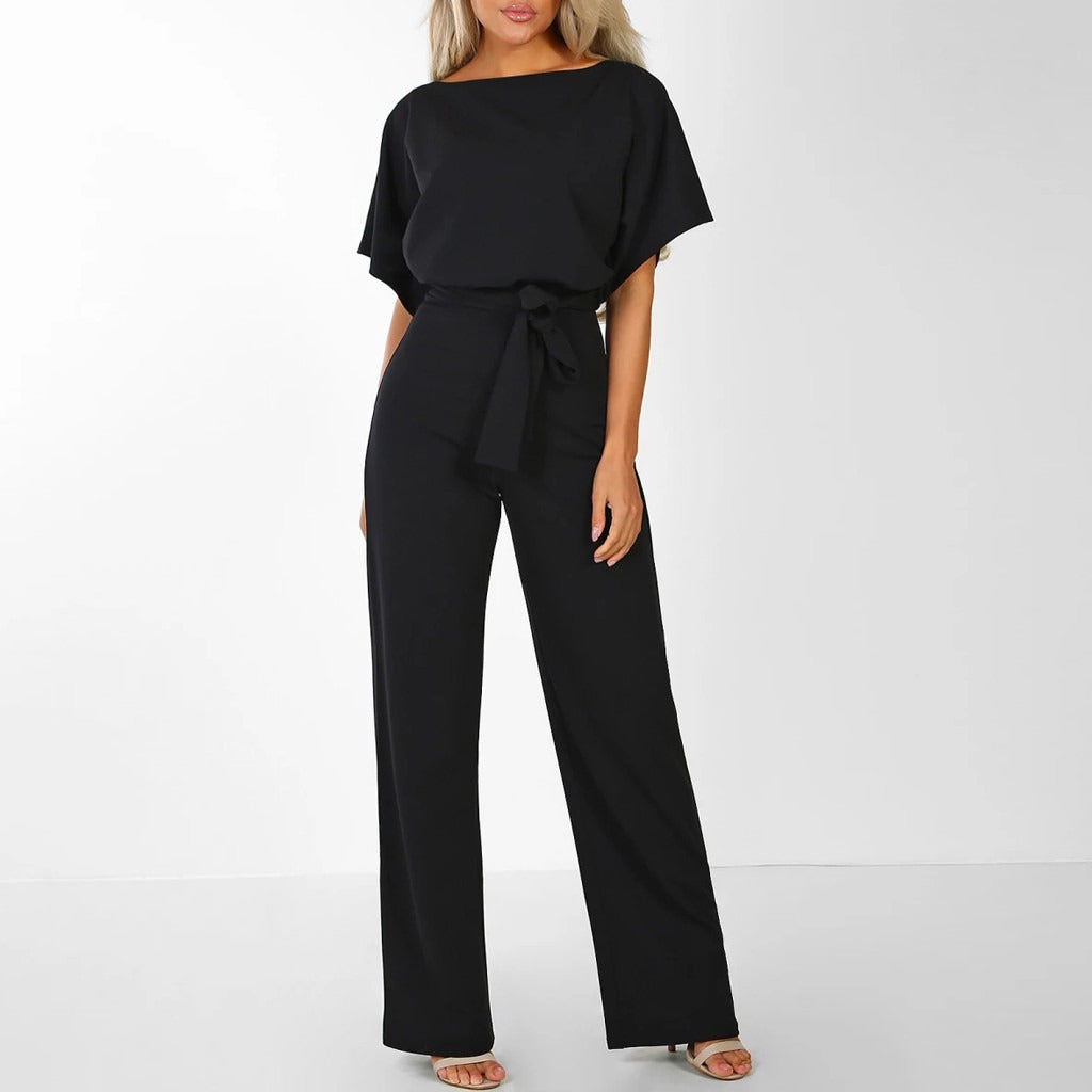Frau in schwarzem Jumpsuit mit Gürtel, weiten Hosenbeinen und kurzen Ärmeln, steht vor weißem Hintergrund. Elegante Damenmode, stilvoller Einteiler.