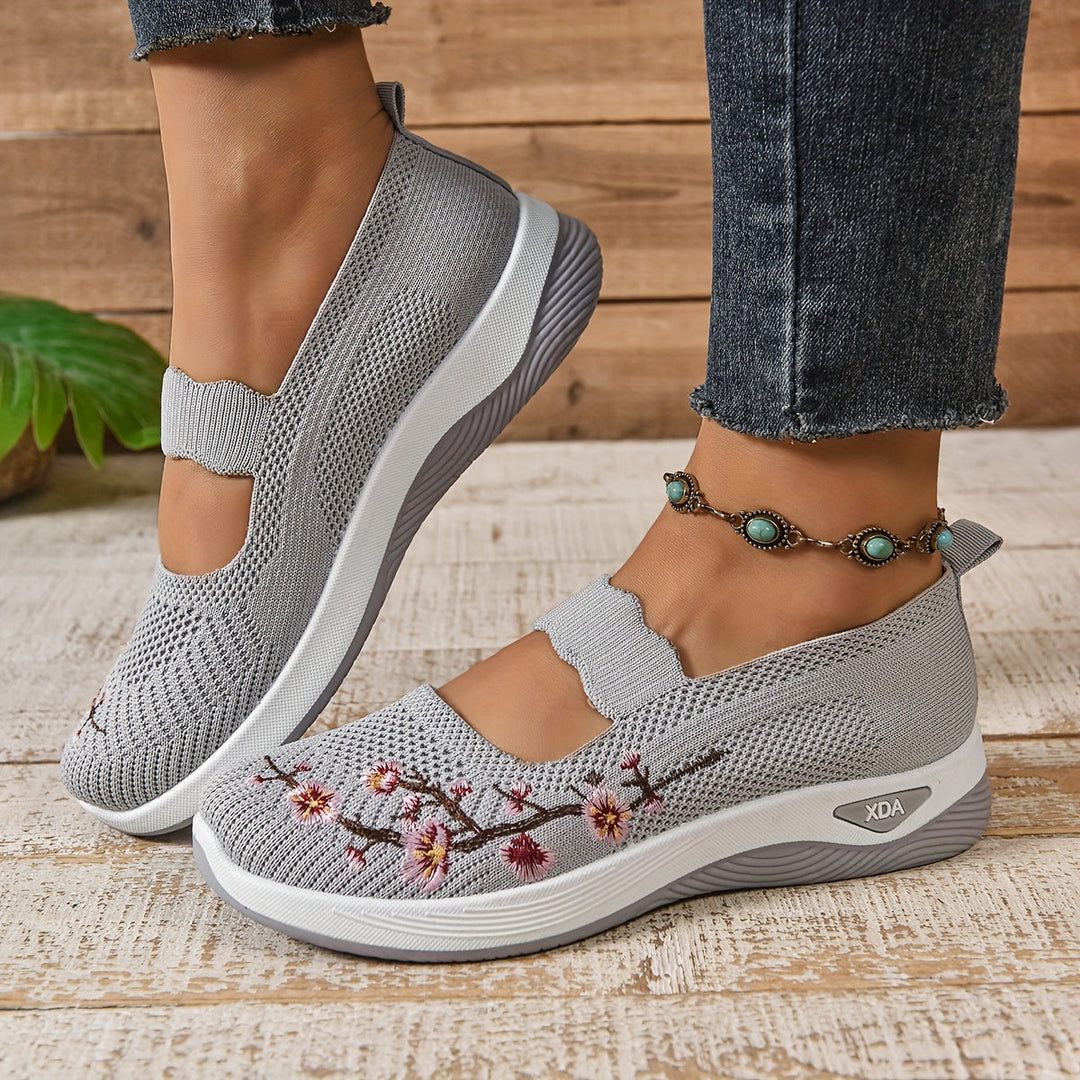 Graue Damen-Sneaker mit Blumenstickerei, atmungsaktivem Mesh-Obermaterial und Keilabsatz, getragen mit Jeans und türkisfarbenem Fußkettchen.