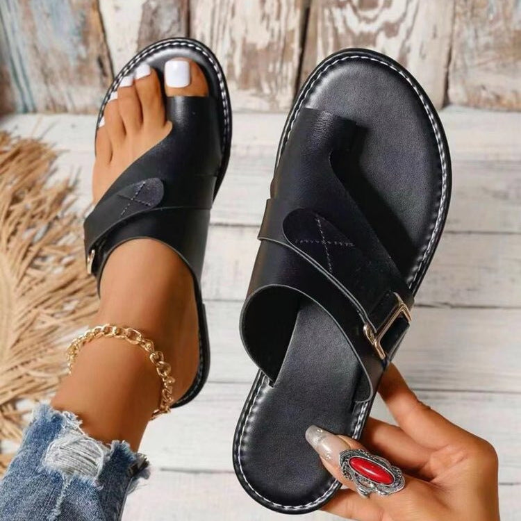 Schwarze Ledersandalen mit Zehenschlaufe, getragen von einer Person mit Jeans und Schmuck. Modische Sommerschuhe auf Holzuntergrund.