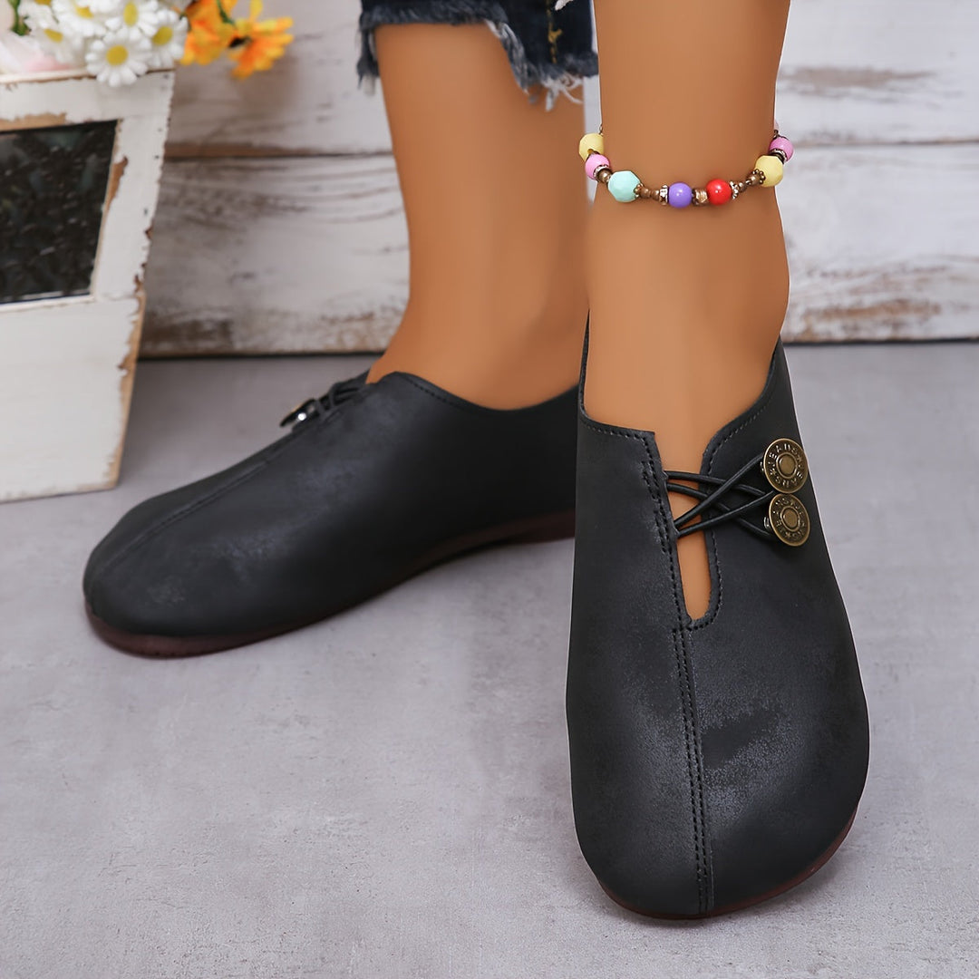 Schwarze Damen-Loafers mit dekorativen Knöpfen, getragen mit bunter Perlenkette am Knöchel. Modische, bequeme Schuhe auf grauem Hintergrund.