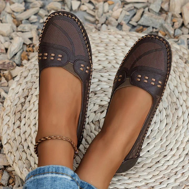 Braune Damen-Loafer aus Leder mit Ziernähten, getragen auf einem geflochtenen Teppich, umgeben von Kieselsteinen. Elegante, bequeme Freizeitschuhe.