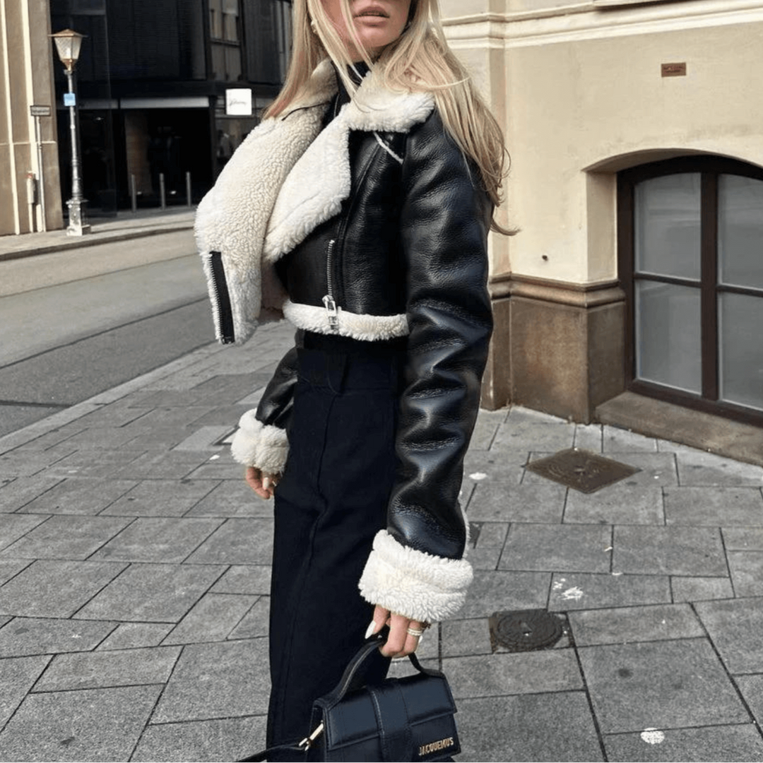 Blonde Frau in schwarzer Lederjacke mit weißem Fellkragen, Sonnenbrille und Handtasche, steht auf städtischer Straße vor modernen Gebäuden.