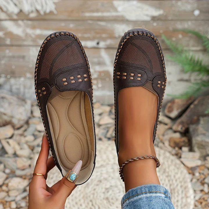 Braune Damen-Loafer aus Leder mit Lochmuster, handgehalten vor rustikalem Hintergrund. Bequeme, stilvolle Schuhe für Freizeit und Alltag.
