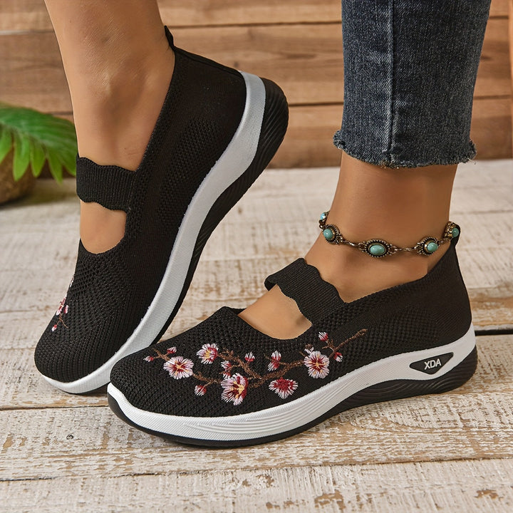 Schwarze Damen-Sneaker mit Blumenstickerei, bequeme Slip-On-Schuhe, kombiniert mit Jeans und türkisfarbenem Fußkettchen auf Holzboden.