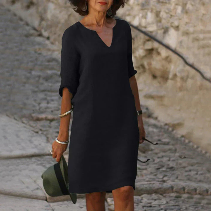 Ältere Frau in elegantem, schwarzem Kleid auf Kopfsteinpflasterstraße, hält Hut in der Hand. Mode für reife Frauen, stilvolles Sommeroutfit.
