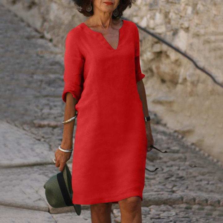 Elegante Frau in rotem Kleid auf Kopfsteinpflasterstraße, hält Hut in der Hand. Modetrend, Sommerkleid, stilvolle Damenmode.