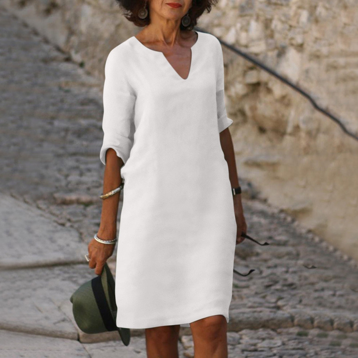 Frau in weißem Sommerkleid auf Kopfsteinpflasterstraße, hält Hut und Sonnenbrille. Elegante Mode, lässiger Stil, urbane Umgebung.