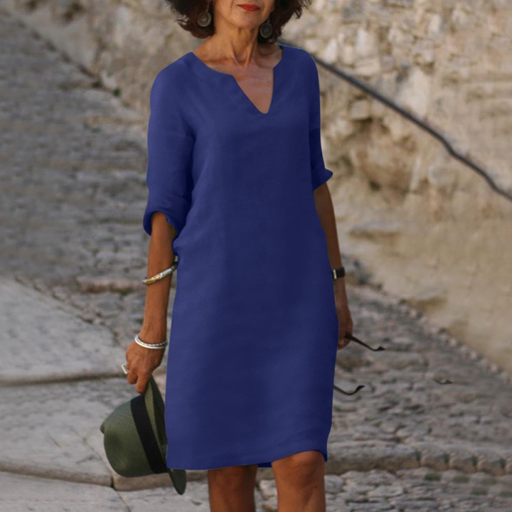 Frau in elegantem, blauem Kleid auf Kopfsteinpflasterstraße, hält Hut in der Hand. Modetrend, Sommerkleid, Damenmode, stilvoller Look.
