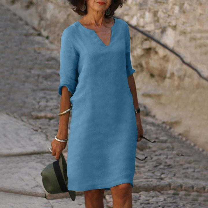 Frau in blauem Sommerkleid mit V-Ausschnitt, hält Hut, geht auf gepflastertem Weg. Elegante Mode, Freizeitkleidung, stilvoller Look.