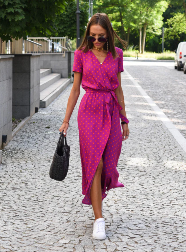 Frau in pinkem Wickelkleid mit Punkten, trägt Sonnenbrille und schwarze Tasche, geht auf gepflastertem Weg. Sommermode, lässiger Stil.