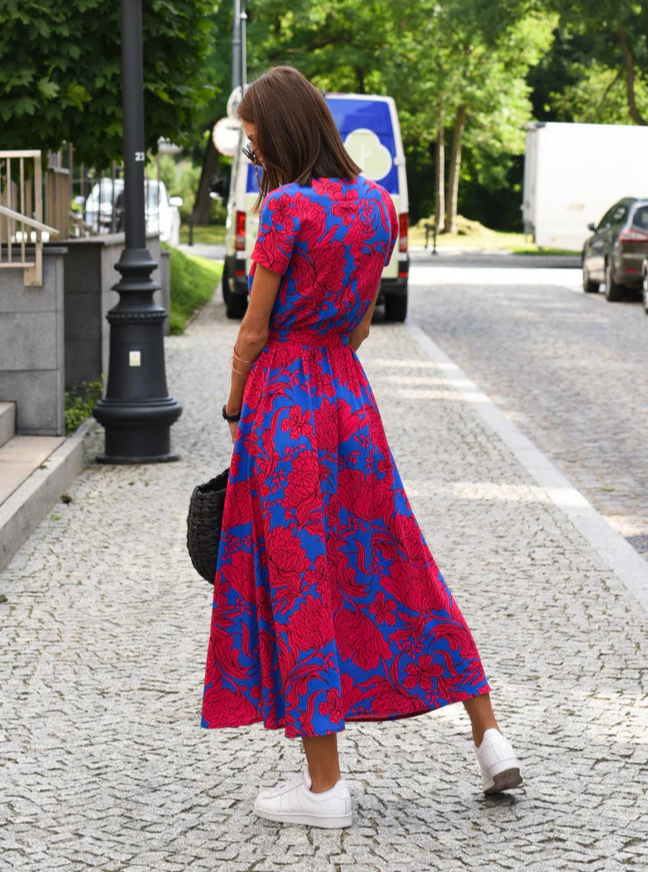 Frau in rotem, geblümtem Kleid auf gepflasterter Straße, trägt weiße Sneaker und schwarze Tasche. Sommermode, Stadtspaziergang, stilvoll.