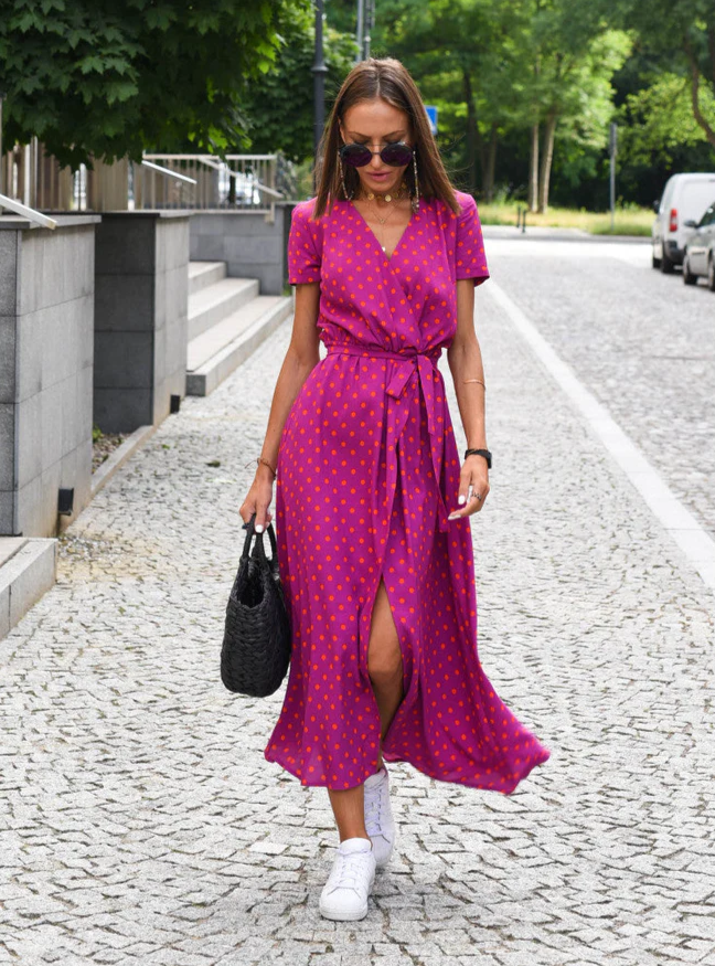 Frau in pinkem, gepunktetem Kleid mit weißen Sneakers und schwarzer Tasche auf gepflasterter Straße. Sommermode, Streetstyle, Damenbekleidung.