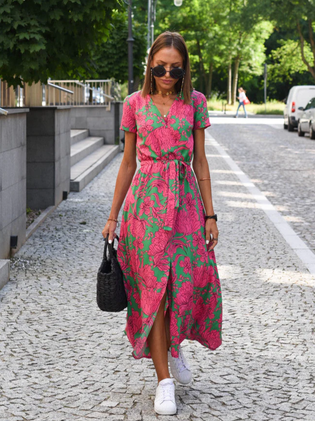 Frau in einem pink-grünen Blumenkleid spaziert auf einer gepflasterten Straße, trägt Sonnenbrille und weiße Sneaker, hält eine schwarze Tasche.