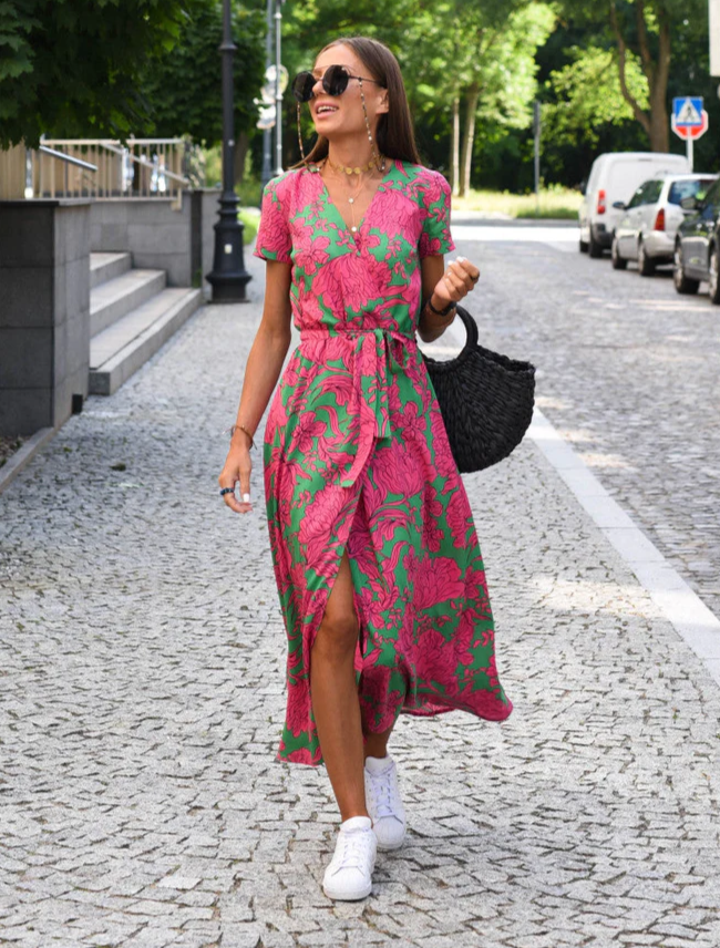 Frau in rosa-grünem Blumenkleid spaziert auf gepflasterter Straße, trägt Sonnenbrille und weiße Sneaker, hält schwarzen Korb. Sommermode, Stadtbummel.