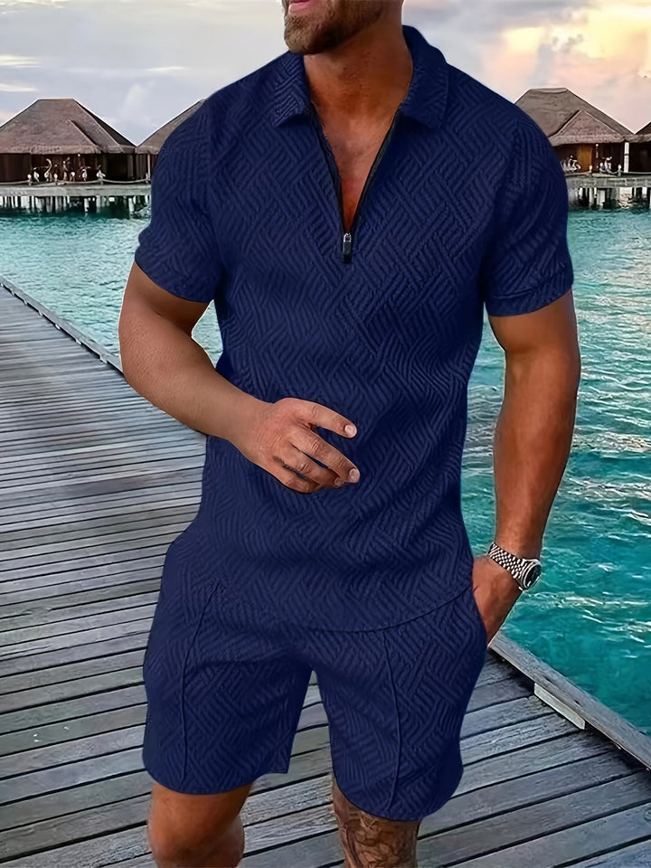 Mann in dunkelblauem, gemustertem Sommeranzug auf Holzsteg am Meer, im Hintergrund Wasserbungalows und Sonnenuntergang.