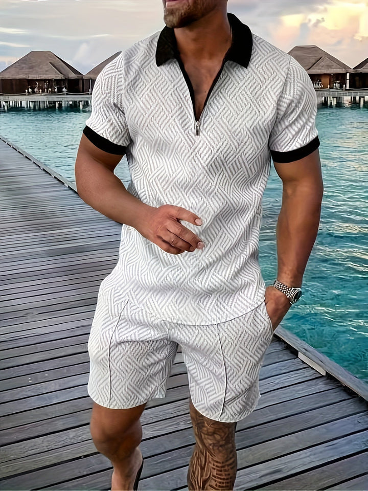 Mann in stilvollem, weißem Sommeroutfit mit geometrischem Muster auf Holzsteg am Meer, im Hintergrund Wasserbungalows, tropische Urlaubsatmosphäre.