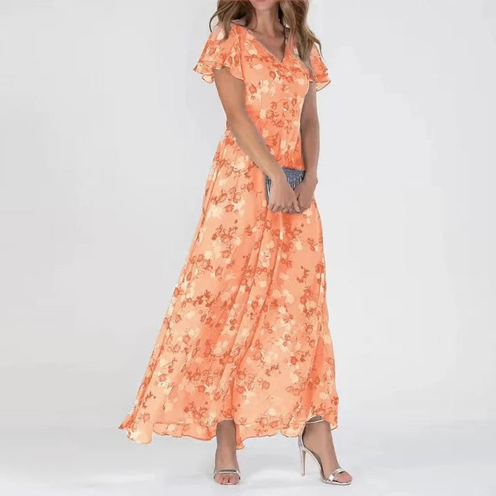 Frau in langem, orangefarbenem Blumenkleid mit kurzen Ärmeln, hält eine graue Clutch, trägt silberne High Heels. Elegantes Sommeroutfit.