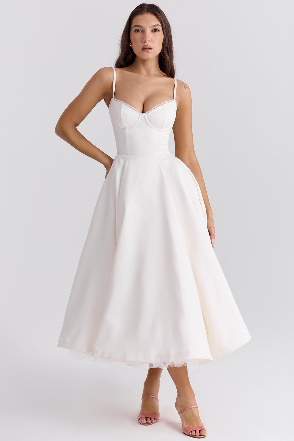 Frau in elegantem, weißem Abendkleid mit Spaghettiträgern und ausgestelltem Rock, stehend vor neutralem Hintergrund.