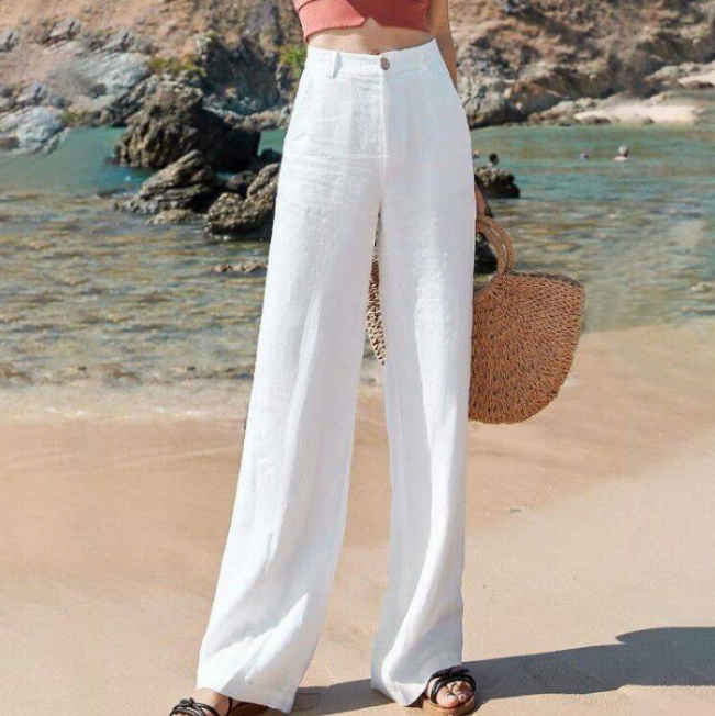 Frau am Strand in weißer Leinenhose, hält Strohtasche. Sommermode, Strandkleidung, lässiger Stil, Urlaubsoutfit.