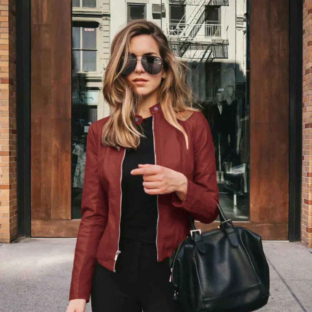 Frau in roter Lederjacke und Sonnenbrille, hält schwarze Handtasche, steht vor einem Gebäude mit Glastüren. Mode, Streetstyle, urbane Eleganz.