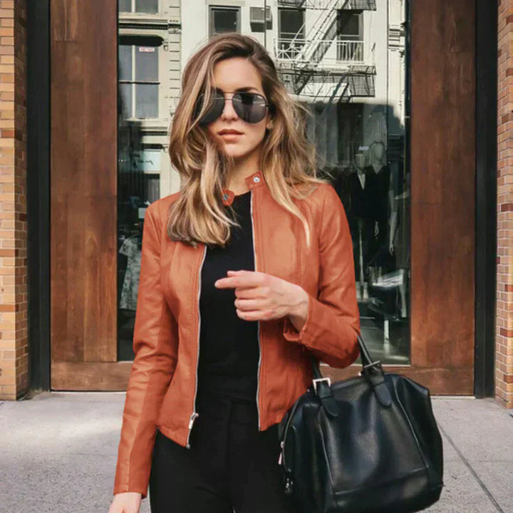 Frau in brauner Lederjacke und Sonnenbrille, hält schwarze Handtasche, steht vor modernem Gebäude. Streetstyle, Mode, urbane Eleganz.