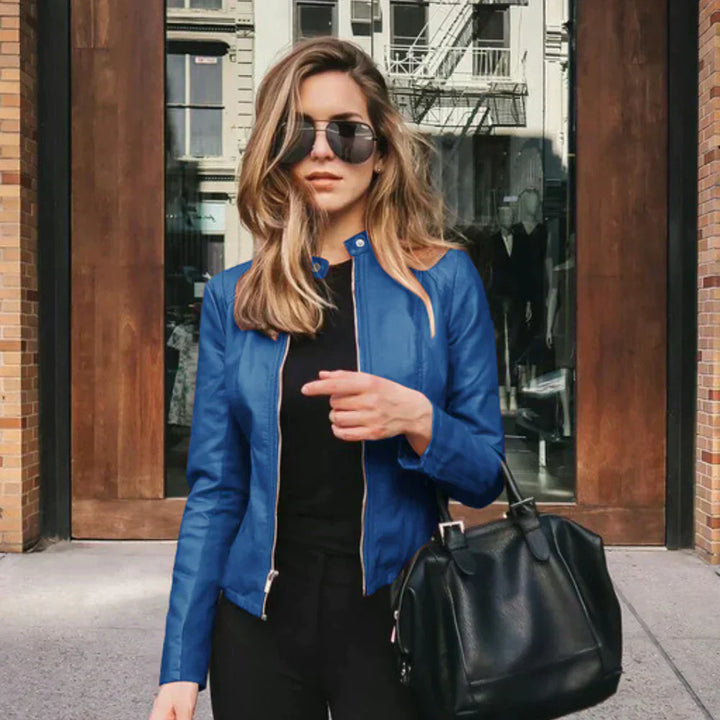 Frau in blauer Lederjacke und Sonnenbrille vor städtischer Kulisse, hält schwarze Handtasche. Modetrend, Streetstyle, urbane Mode.