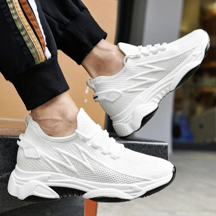 Weiße Sportschuhe mit atmungsaktivem Mesh-Obermaterial und rutschfester Sohle, getragen von einer Person in schwarzer Hose mit Streifen.
