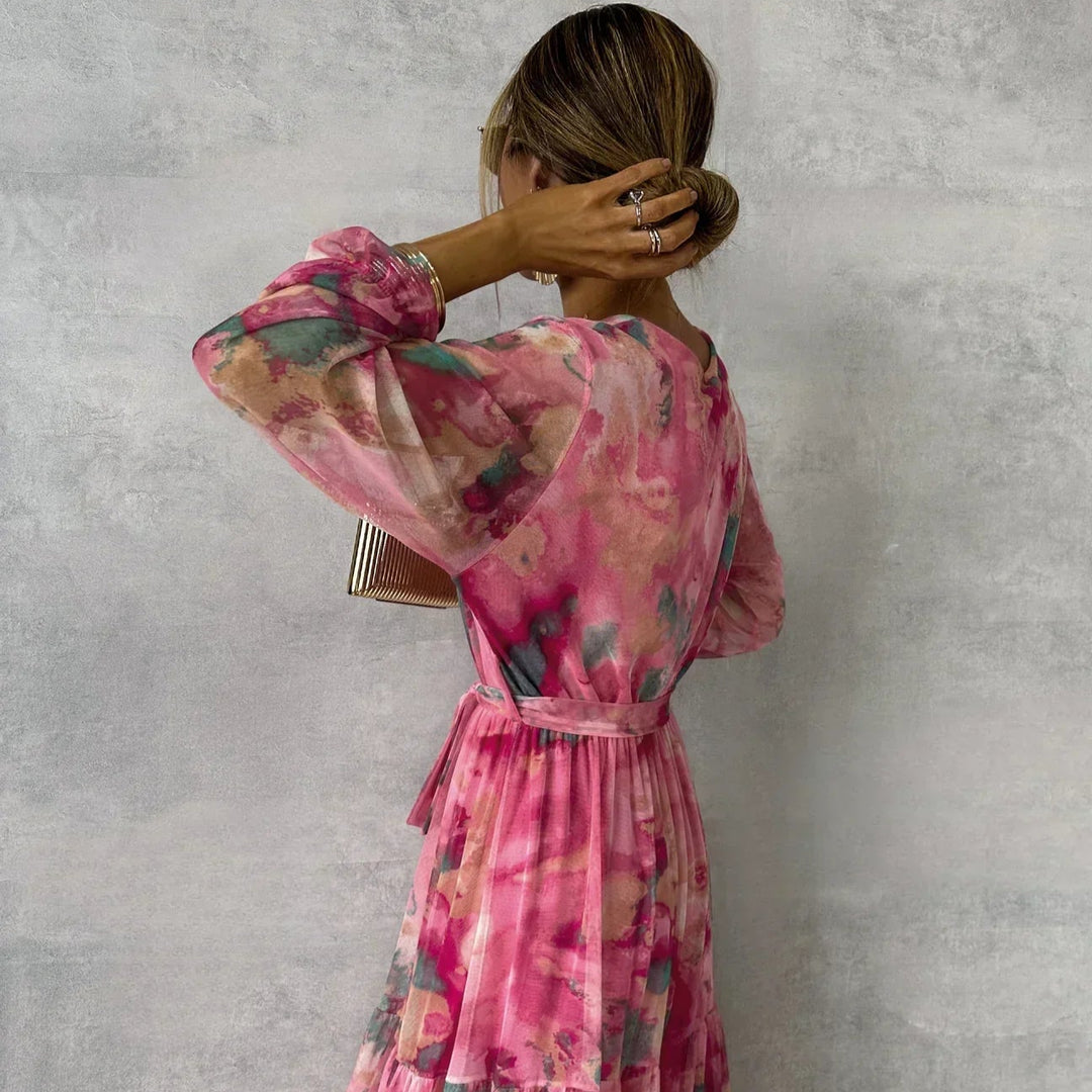 Frau in langem, geblümtem Kleid in Rosa- und Grüntönen, seitlich fotografiert, hält eine kleine Tasche. Elegante Mode, Sommerkleid, florales Design.