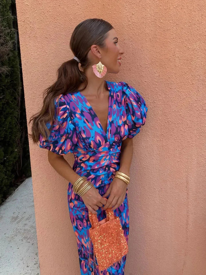 Frau in blauem, gemustertem Kleid mit Puffärmeln, orangefarbene Ohrringe und Tasche, steht vor pfirsichfarbener Wand, modisches Sommeroutfit.