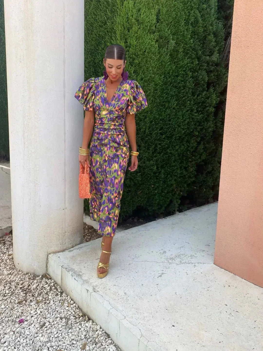 Frau in buntem, gemustertem Kleid mit Puffärmeln, hält orangefarbene Tasche, steht vor grüner Hecke und Säule. Sommermode, eleganter Look.