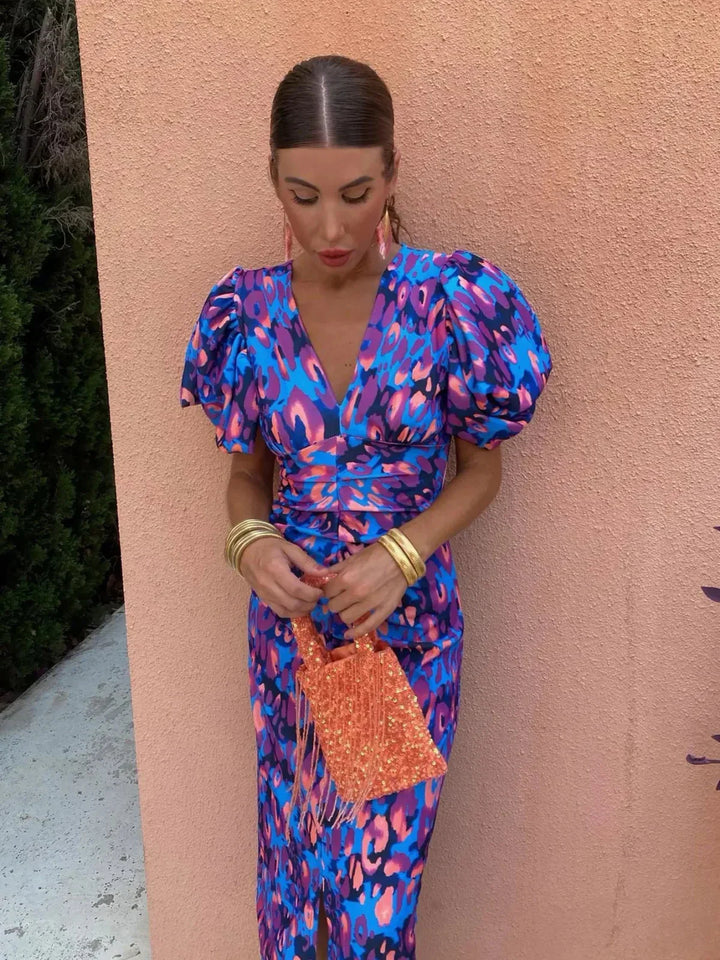 Frau in buntem Kleid mit Puffärmeln und orangefarbener Tasche vor rosa Wand. Modetrend, Sommeroutfit, stilvolle Accessoires.