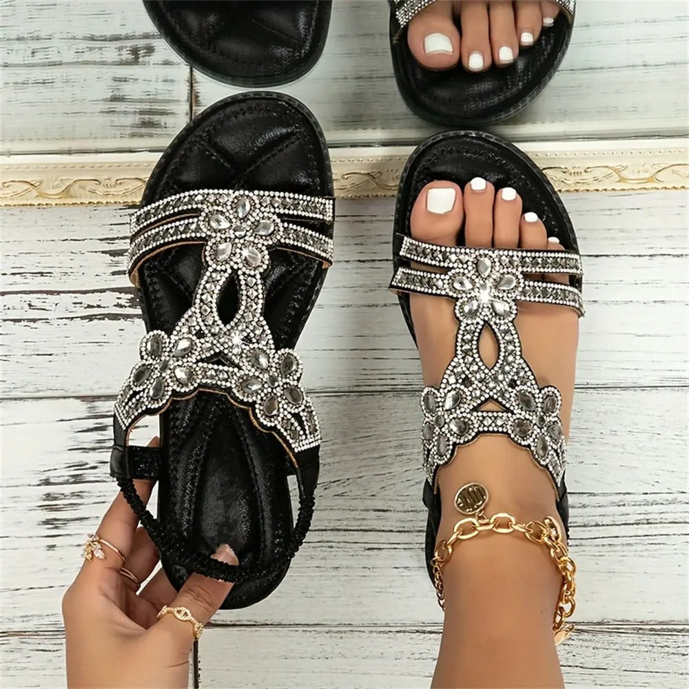 Schwarze Sandalen mit glitzernden Strassblumen, getragen von einer Person mit weiß lackierten Zehennägeln, auf hellem Holzboden.