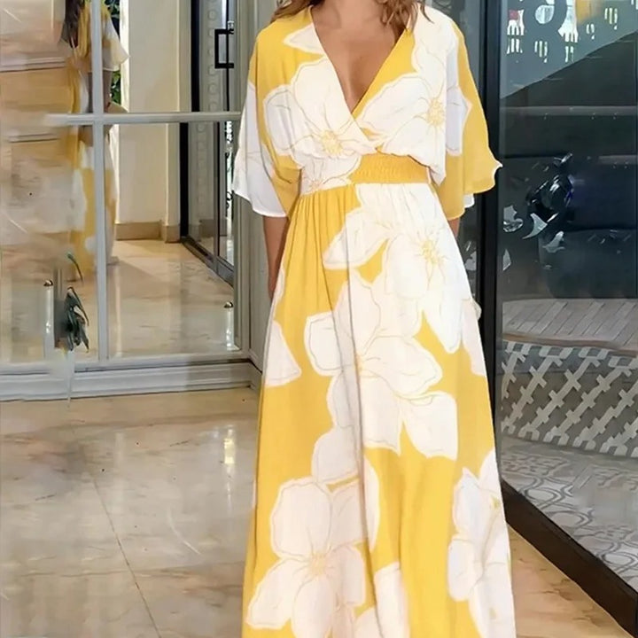 Frau in gelbem Maxikleid mit weißen Blumen, steht in modernem Innenraum. Sommermode, elegantes Design, florales Muster.