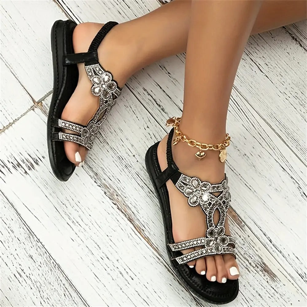 Schwarze Sandalen mit Strasssteinen und Blumenmuster auf weißem Holzboden, Fuß mit goldener Fußkette. Elegante Sommerschuhe für Damen.