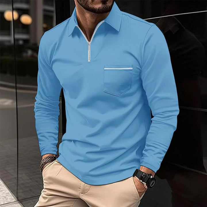 Mann in hellblauem Langarmshirt mit Reißverschluss und Brusttasche, kombiniert mit beigen Hosen. Modischer Freizeitlook, urbaner Hintergrund.