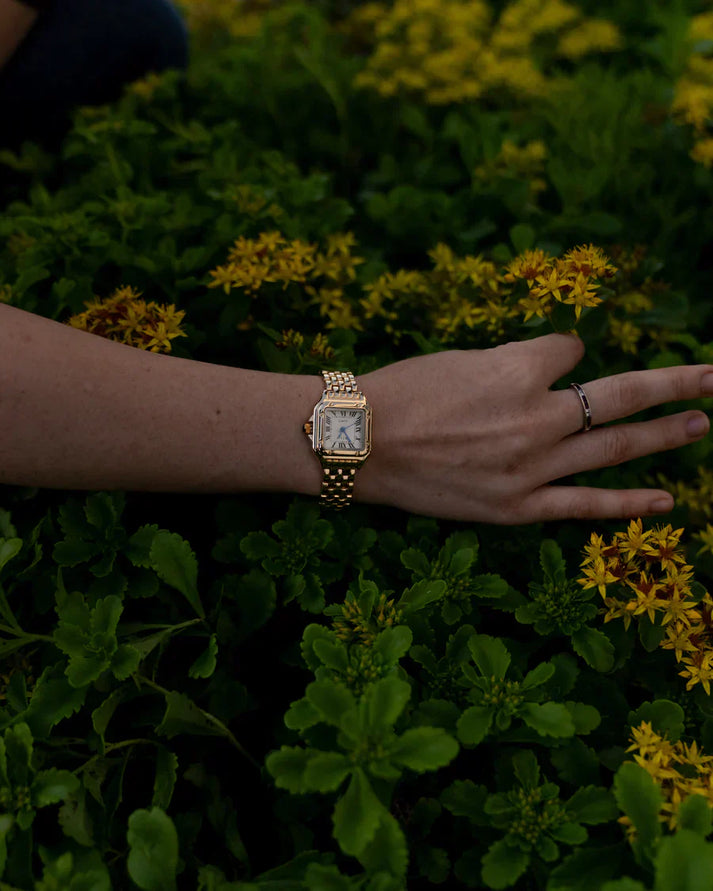 Goldene Armbanduhr an einem Handgelenk, umgeben von gelben Blumen und grünem Laub. Luxuriöse Uhr, Natur, stilvolles Accessoire.