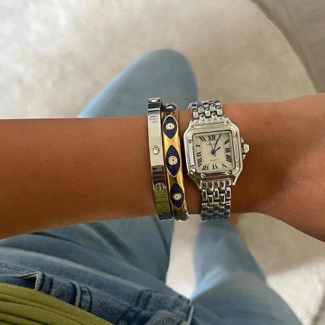 Armbanduhr mit silbernem Gliederarmband und römischen Ziffern, kombiniert mit drei modischen Armreifen in Gold und Blau auf einem Handgelenk.