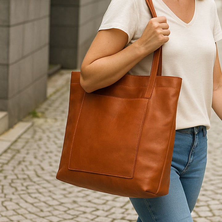 Frau mit brauner Ledertasche, lässig über der Schulter, trägt weißes T-Shirt und Jeans. Stilvolle Handtasche, modisches Accessoire, Stadtumgebung.