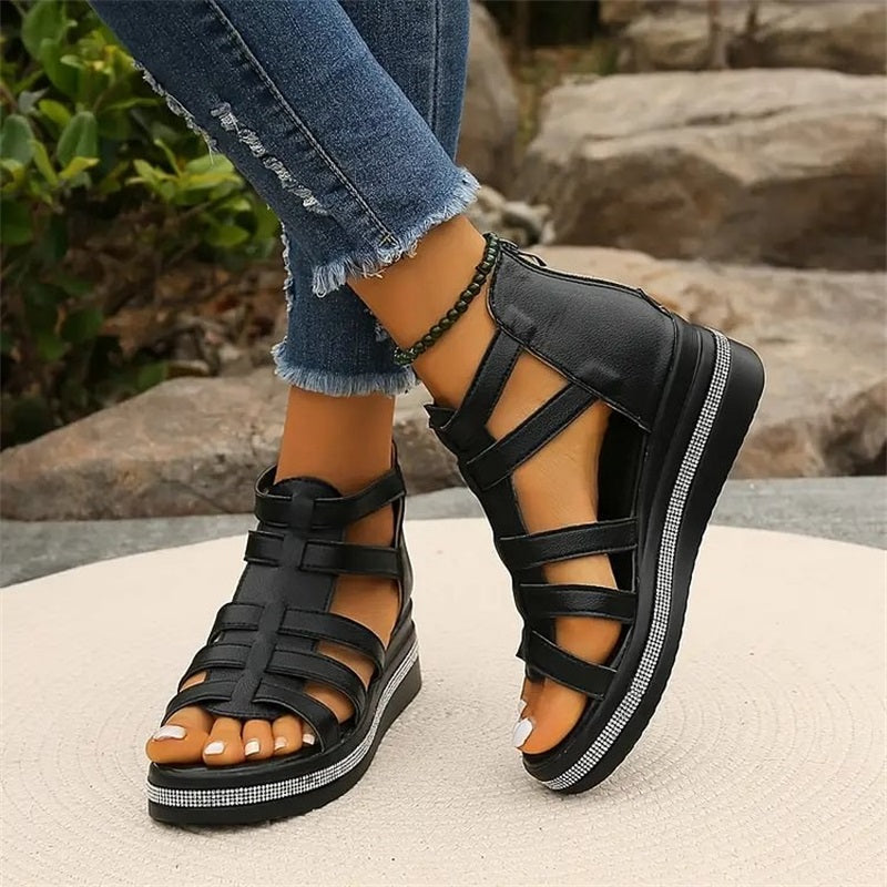 Schwarze Gladiatorensandalen mit Keilabsatz, getragen von einer Person in zerrissenen Jeans, im Freien auf Steinboden. Modische Sommerschuhe Damen.