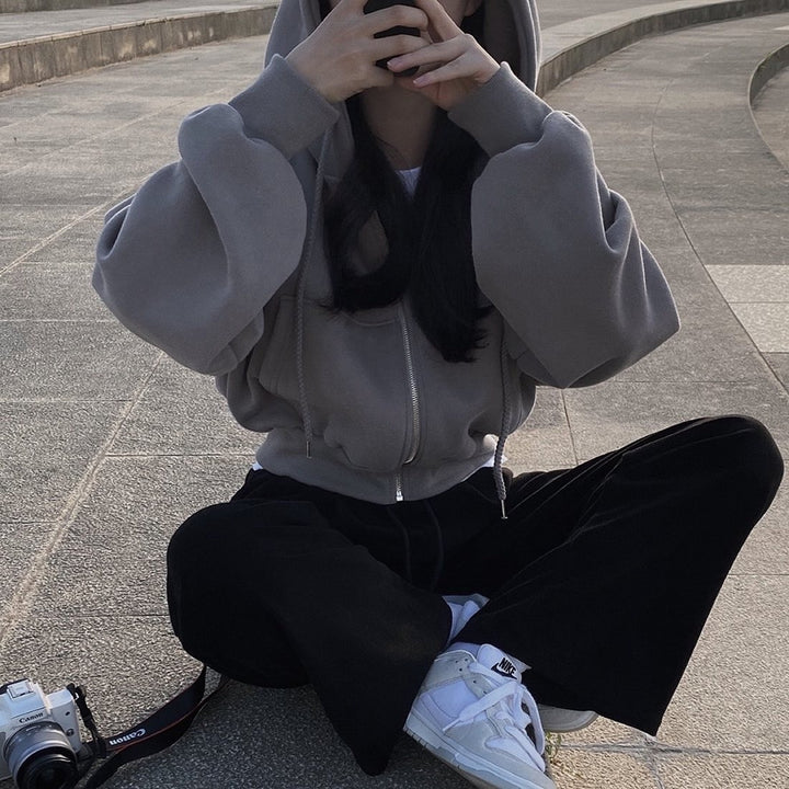 Frau in grauem Hoodie und schwarzen Hosen sitzt im Freien, hält Smartphone, Kamera neben ihr. Freizeitmode, Outdoor, Fotografie.