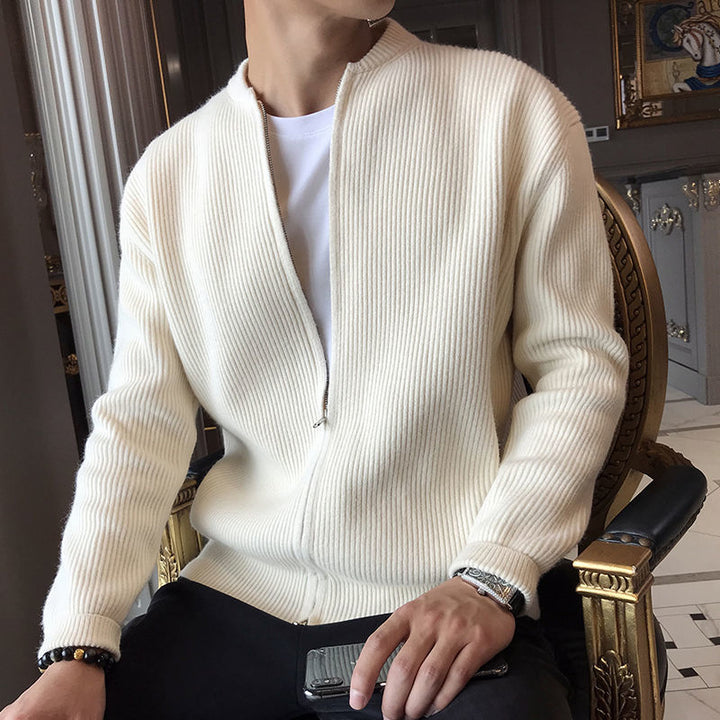 Mann in weißer Strickjacke mit Reißverschluss, sitzt auf einem Stuhl, hält ein Smartphone. Elegante Herrenmode, stilvoller Freizeitlook.